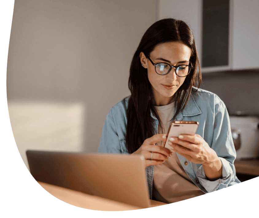 Žena v okuliaroch si prezerá informácie na smartfóne vedľa otvoreného notebooku.